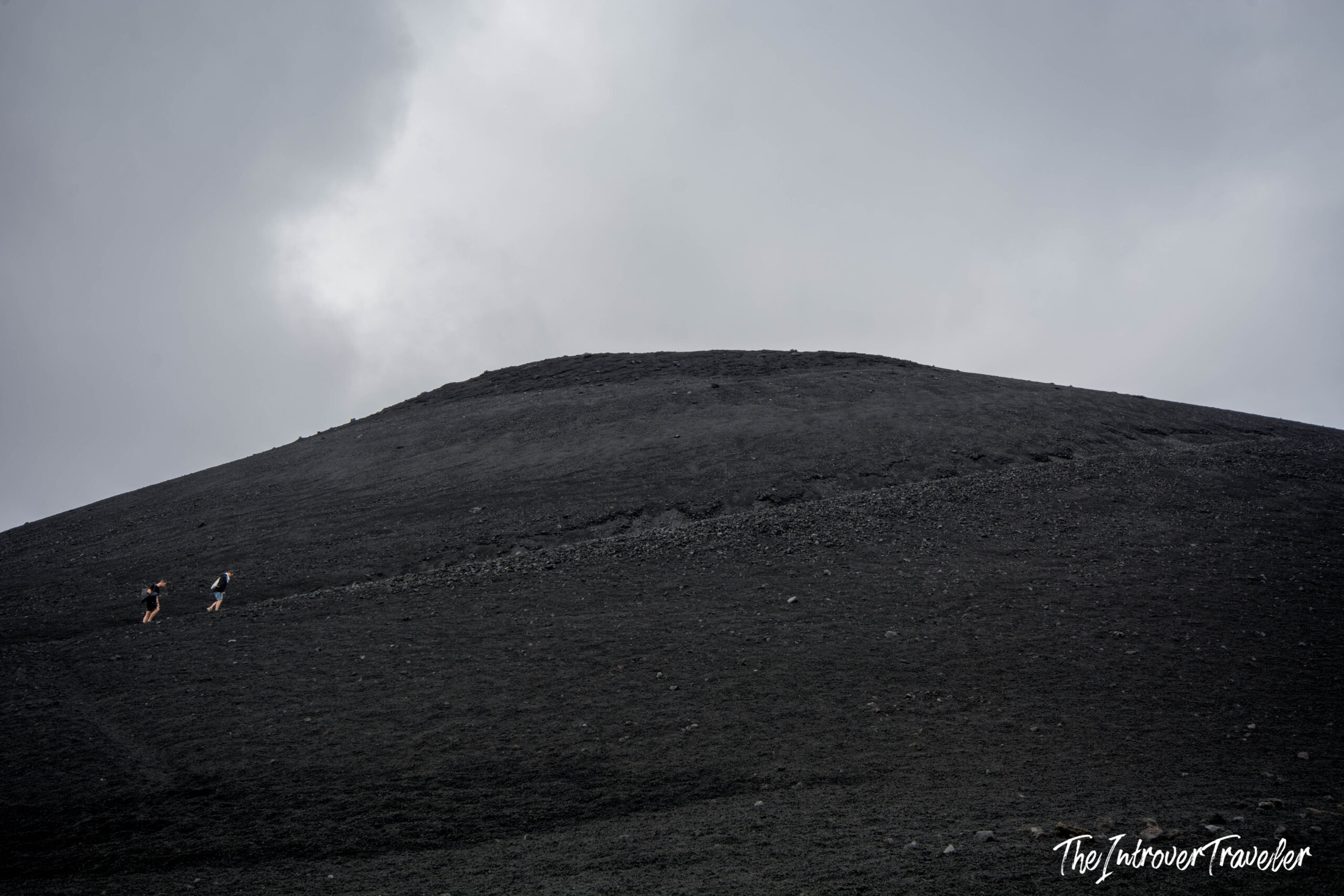 Two hikers ascending the steep black lava slopes of Mount Etna under an overcast sky