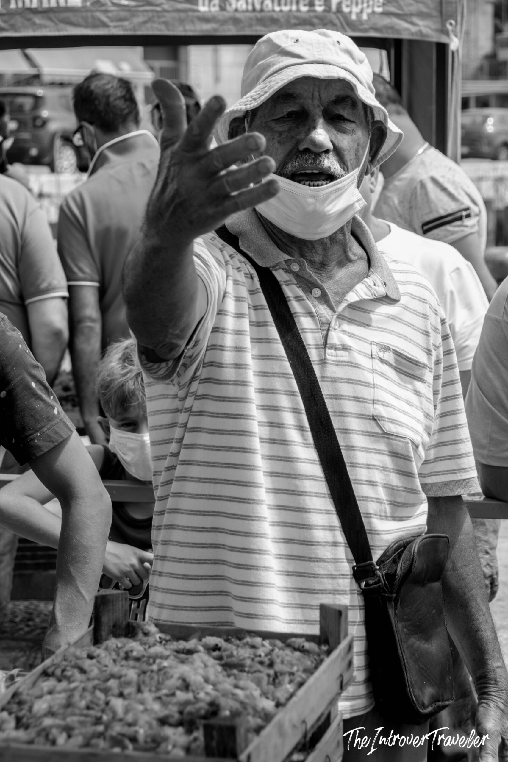 Animated vendor gesturing at a busy coastal fish market stall, mask lowered, bucket hat worn