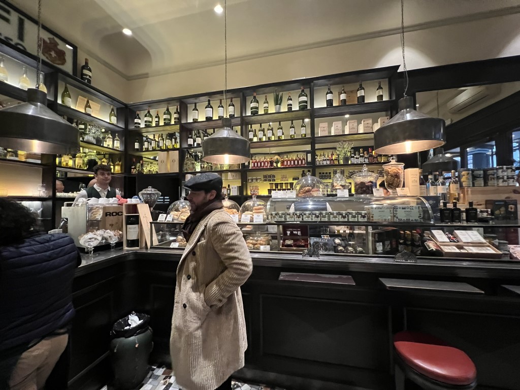 Interior of Procacci 1885 Florence, showing wine shelves, pastry counter, and a customer at the bar