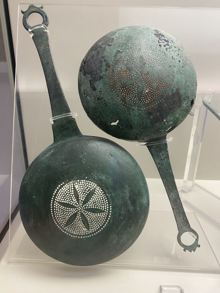 Two ancient Etruscan bronze strainers with pierced geometric patterns, displayed at Villa Giulia museum, Rome