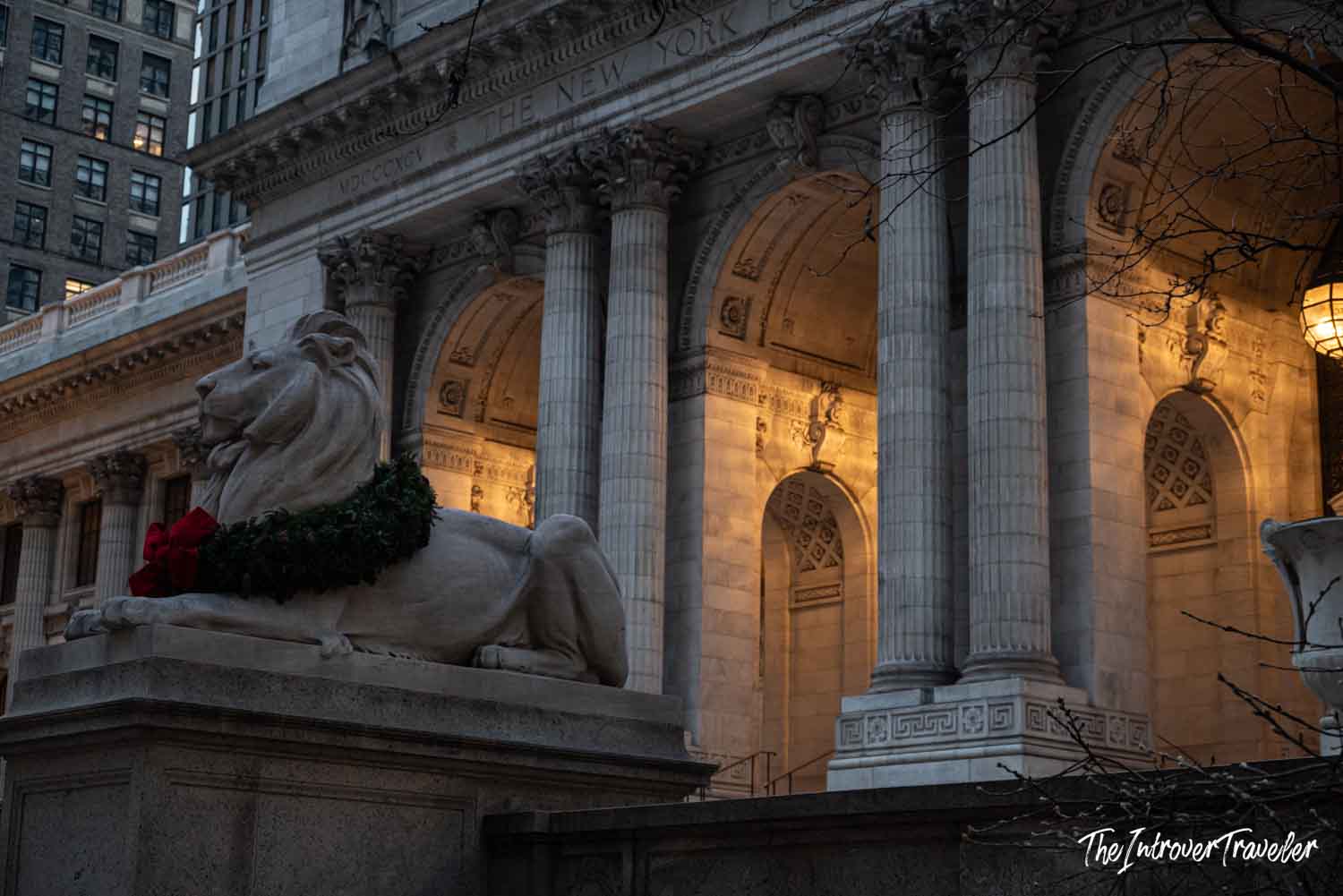 New York Public Library lion statue wearing a Christmas wreath, with the Beaux-Arts facade lit at dusk