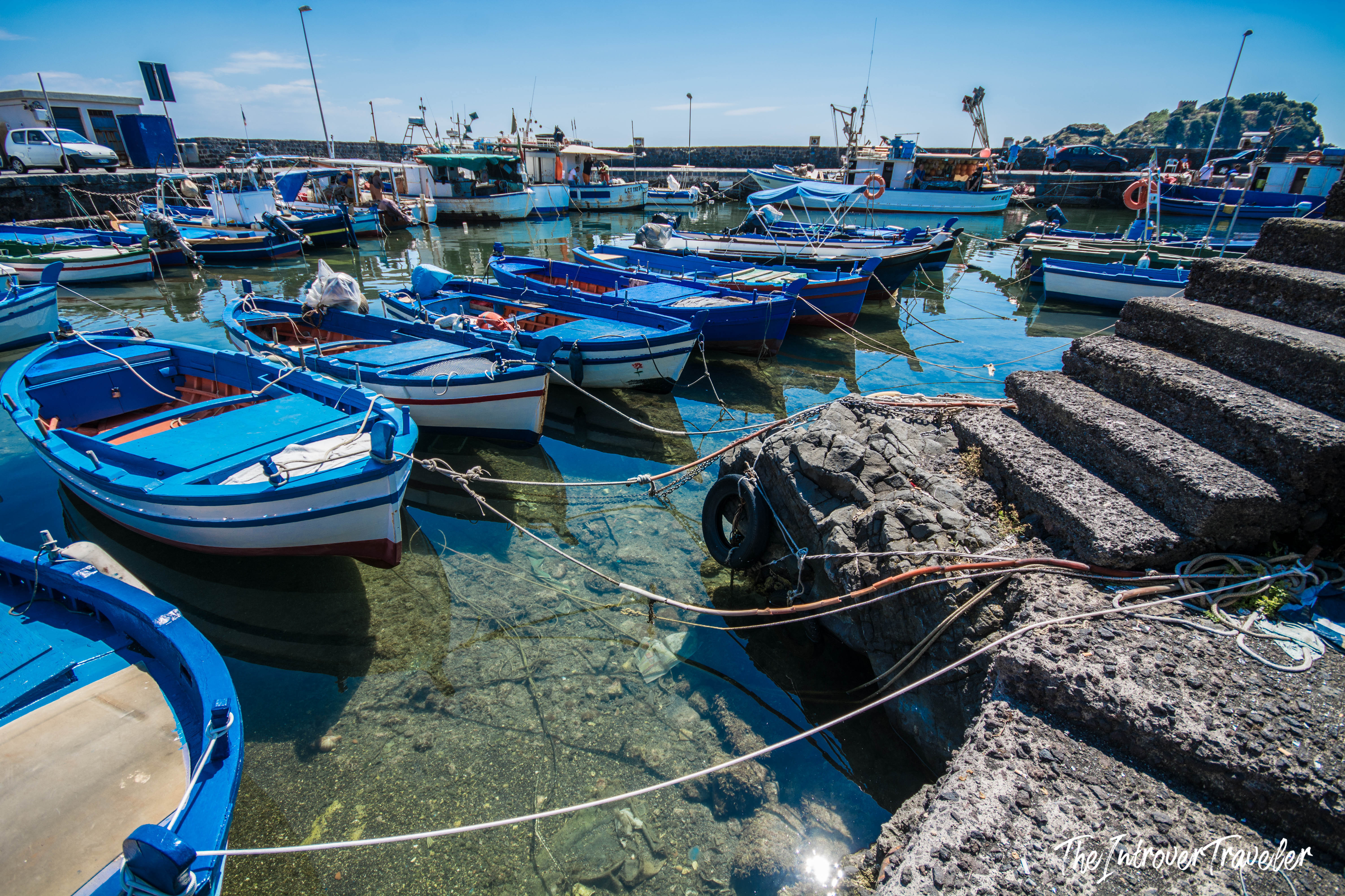 Blue wooden fishing boats moored at Aci Trezza harbor, with volcanic basalt steps and clear Sicilian waters