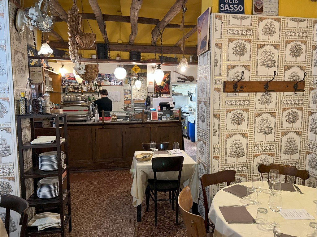 Cozy interior of Trattoria da Maro in Bologna, with rustic beams, patterned wallpaper, and set tables