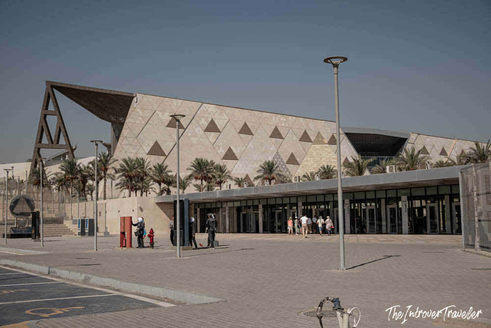 Exterior of the Grand Egyptian Museum in Cairo, with its geometric triangle-patterned facade and palm trees lining the entrance.