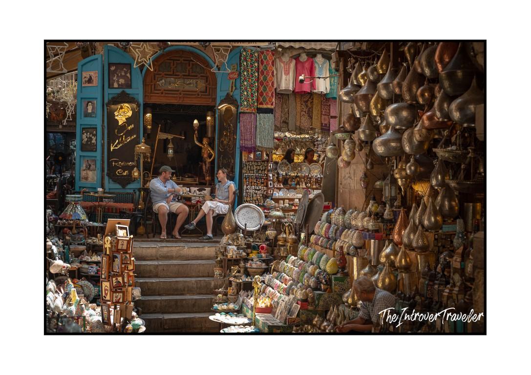 Two tourists resting on steps at Khan el-Khalili bazaar, Cairo, surrounded by brass goods and souvenirs