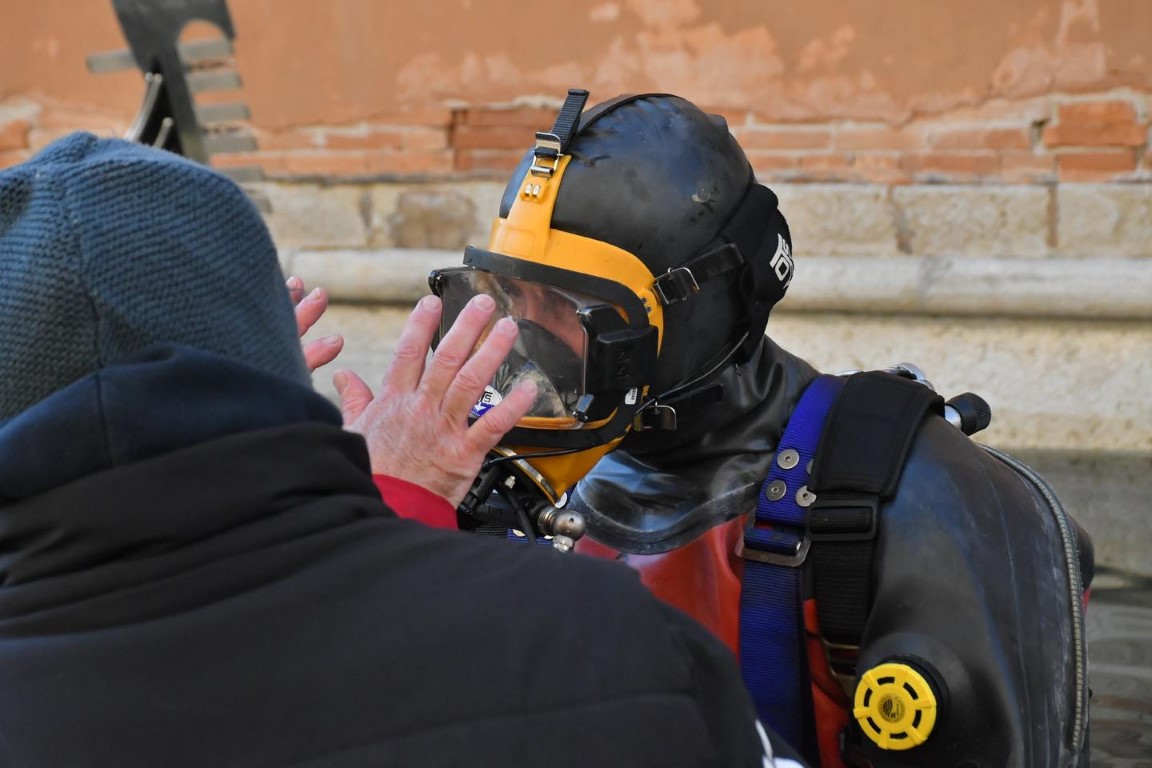 Diver in full wetsuit and mask receiving briefing beside a Venice canal wall