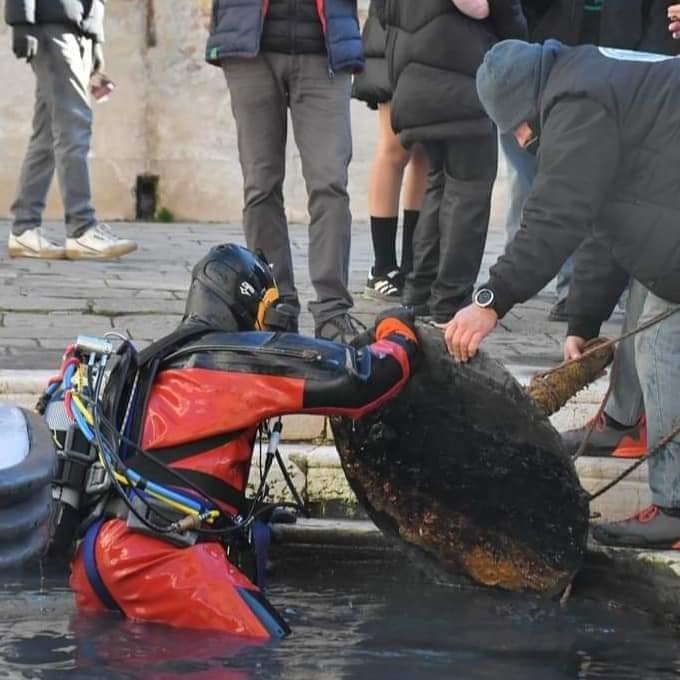 Diver in red drysuit retrieving a large object from a Venice canal, watched by onlookers