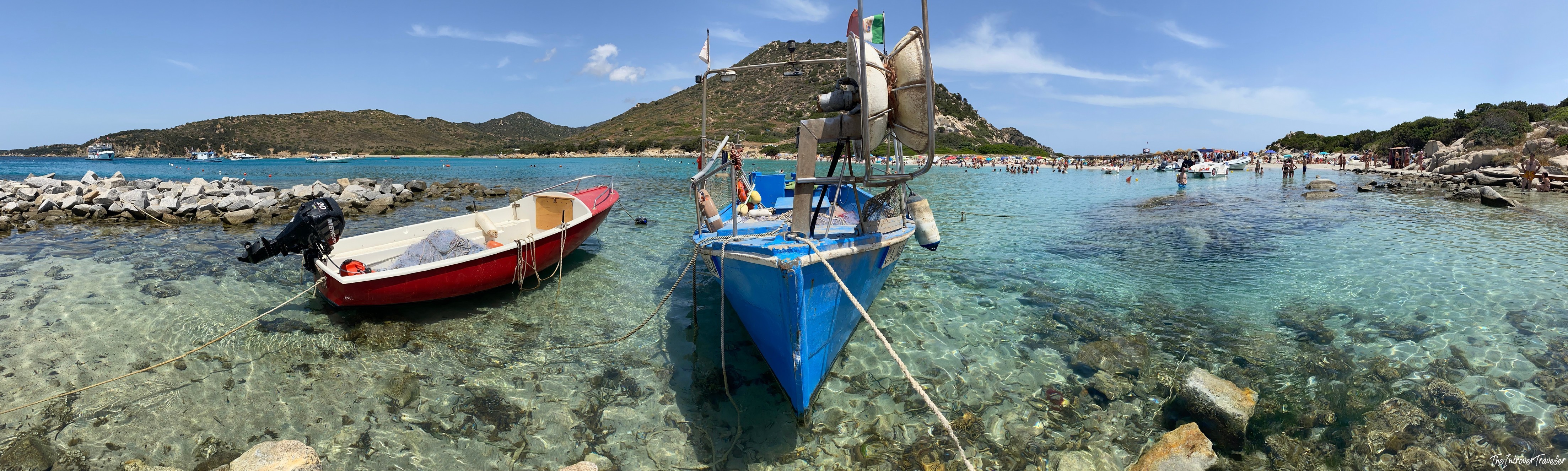 Blue and red fishing boats moored in crystal-clear turquoise water at a busy south Sardinia beach