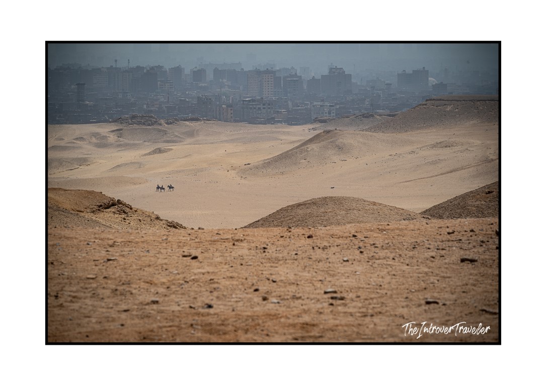 Vast Giza desert plateau with two horse riders dwarfed by sand dunes, Cairo skyline in the haze behind