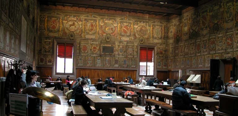 Interior of the Archiginnasio Palace library in Bologna, with heraldic frescoes covering the walls above students reading at wooden desks.