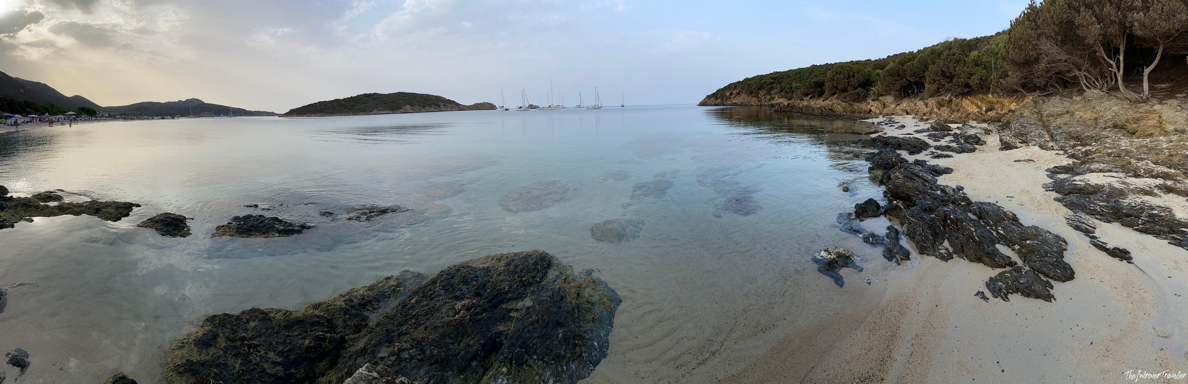 Panoramic view of a sheltered south Sardinia cove with clear turquoise water, dark rocks, and sailboats anchored offshore