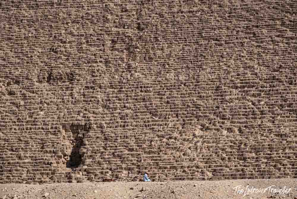 Tiny figure sitting at the base of a Giza pyramid, showing the massive scale of its limestone blocks