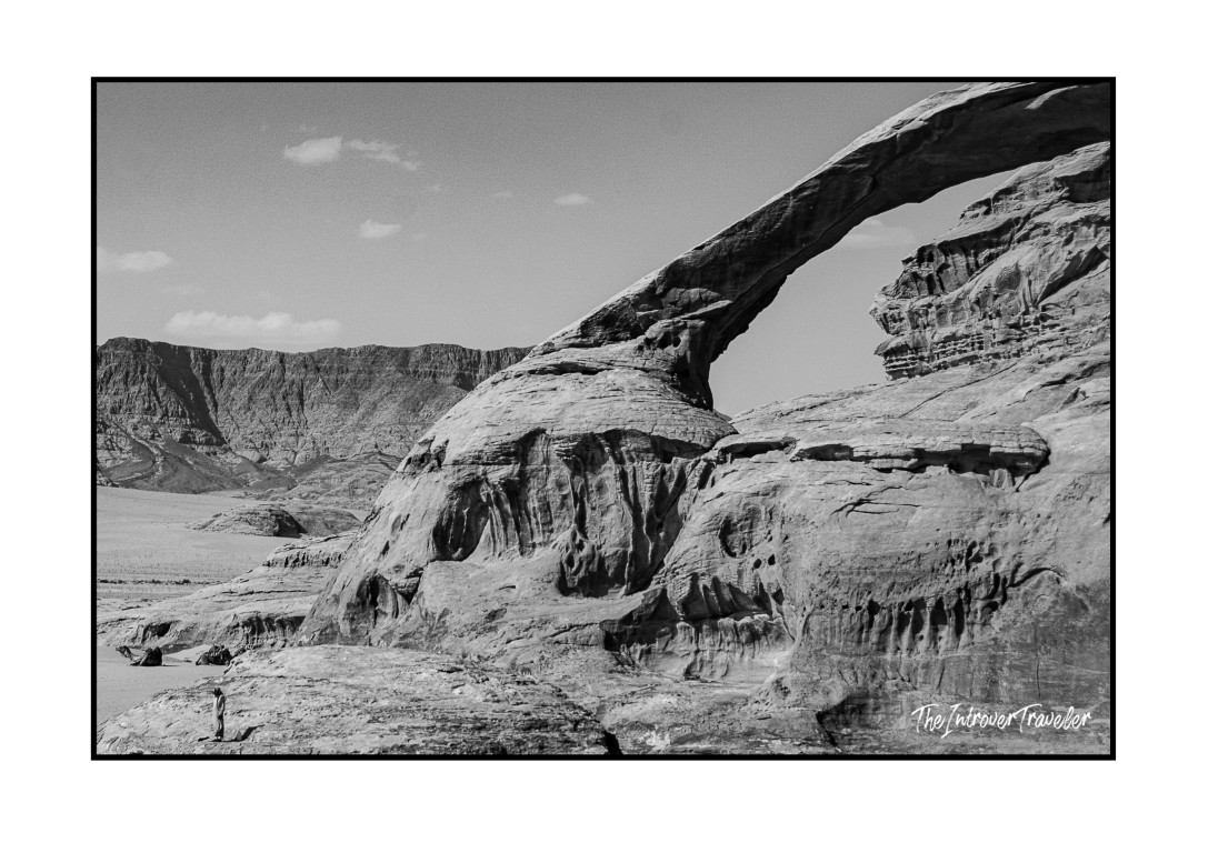 Black and white photo of Burdah Rock Bridge in Wadi Rum, Jordan, with a lone figure showing its massive scale