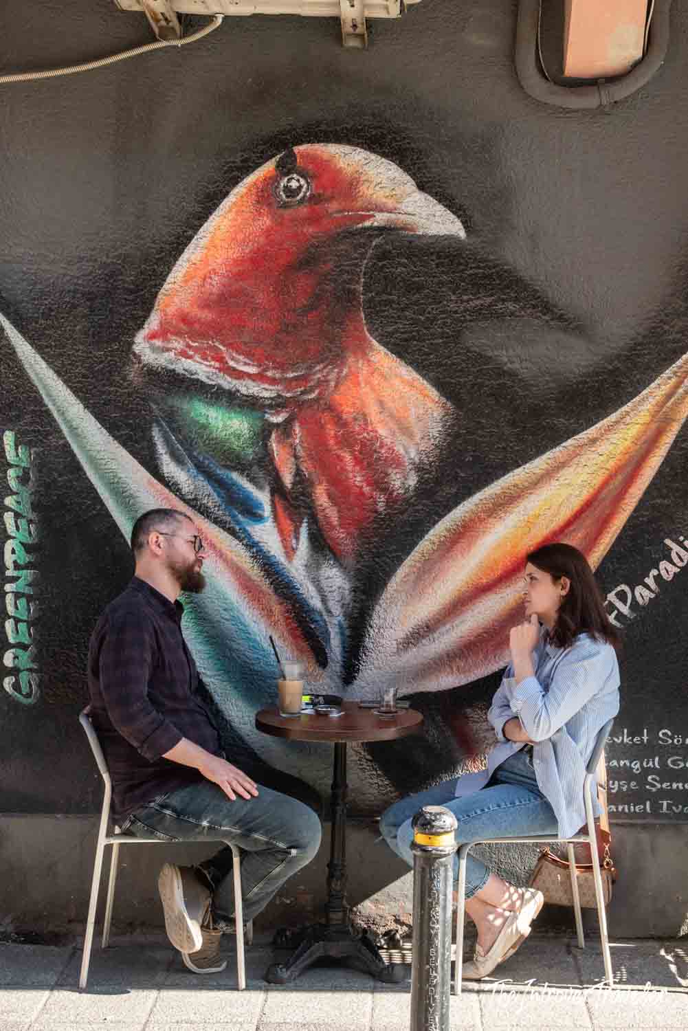 Two people at a cafe table beside a Greenpeace bird of paradise street art mural in Kadıköy, Istanbul