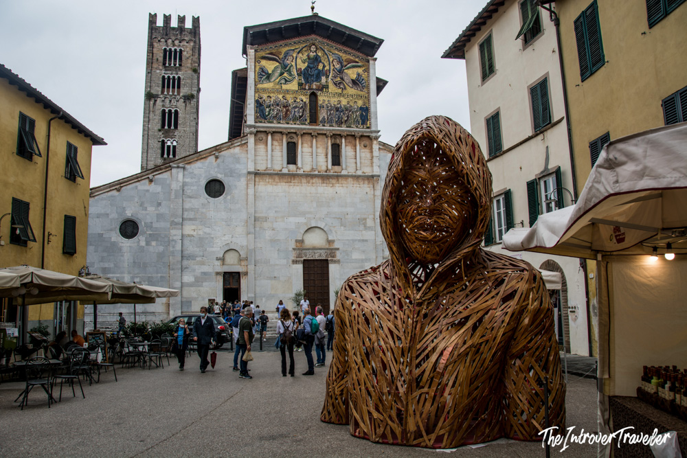 Woven wooden hooded sculpture in Piazza San Frediano, Lucca, with the mosaic basilica facade behind