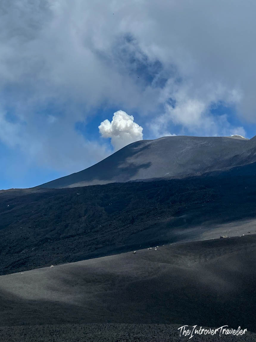 Mount Etna summit crater releasing a white smoke puff above dark volcanic lava fields, Sicily