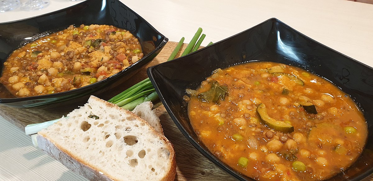 Two black bowls of Tuscan chickpea and lentil stew served with crusty bread and spring onions
