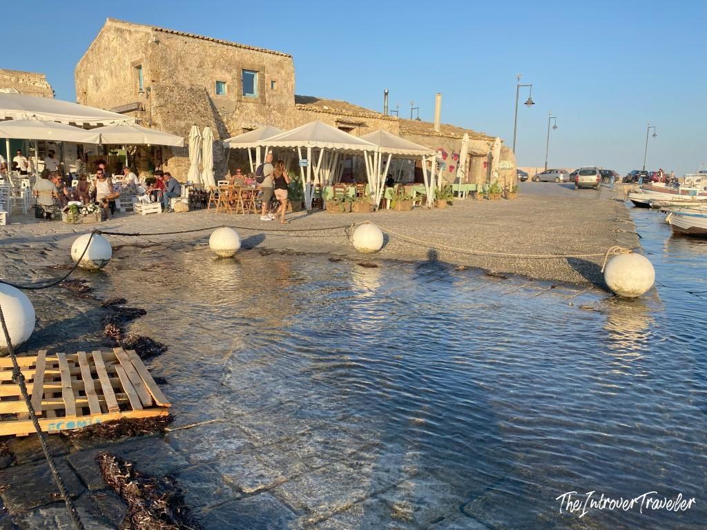 Marzamemi harbor waterfront with outdoor restaurants, stone buildings, and mooring buoys in Sicily