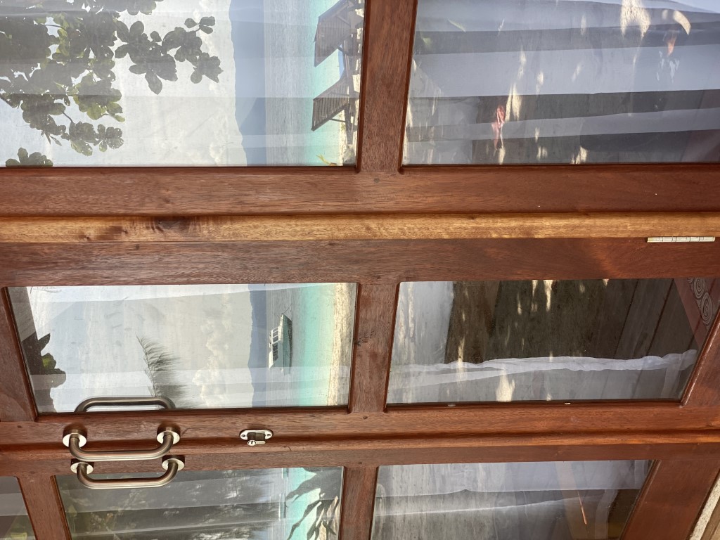 Wooden glass door view of Raja Ampat beach, turquoise water, and a moored boat from a resort bungalow