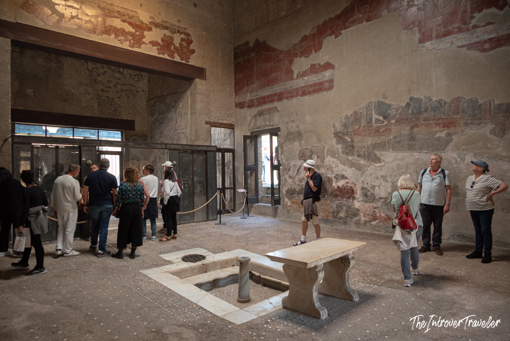 Tourists exploring the atrium of a Herculaneum house, with faded frescoes and a marble impluvium visible