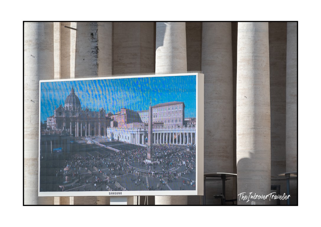 Samsung screen showing aerial view of St. Peter's Square, positioned between Bernini's colonnade columns at the Vatican