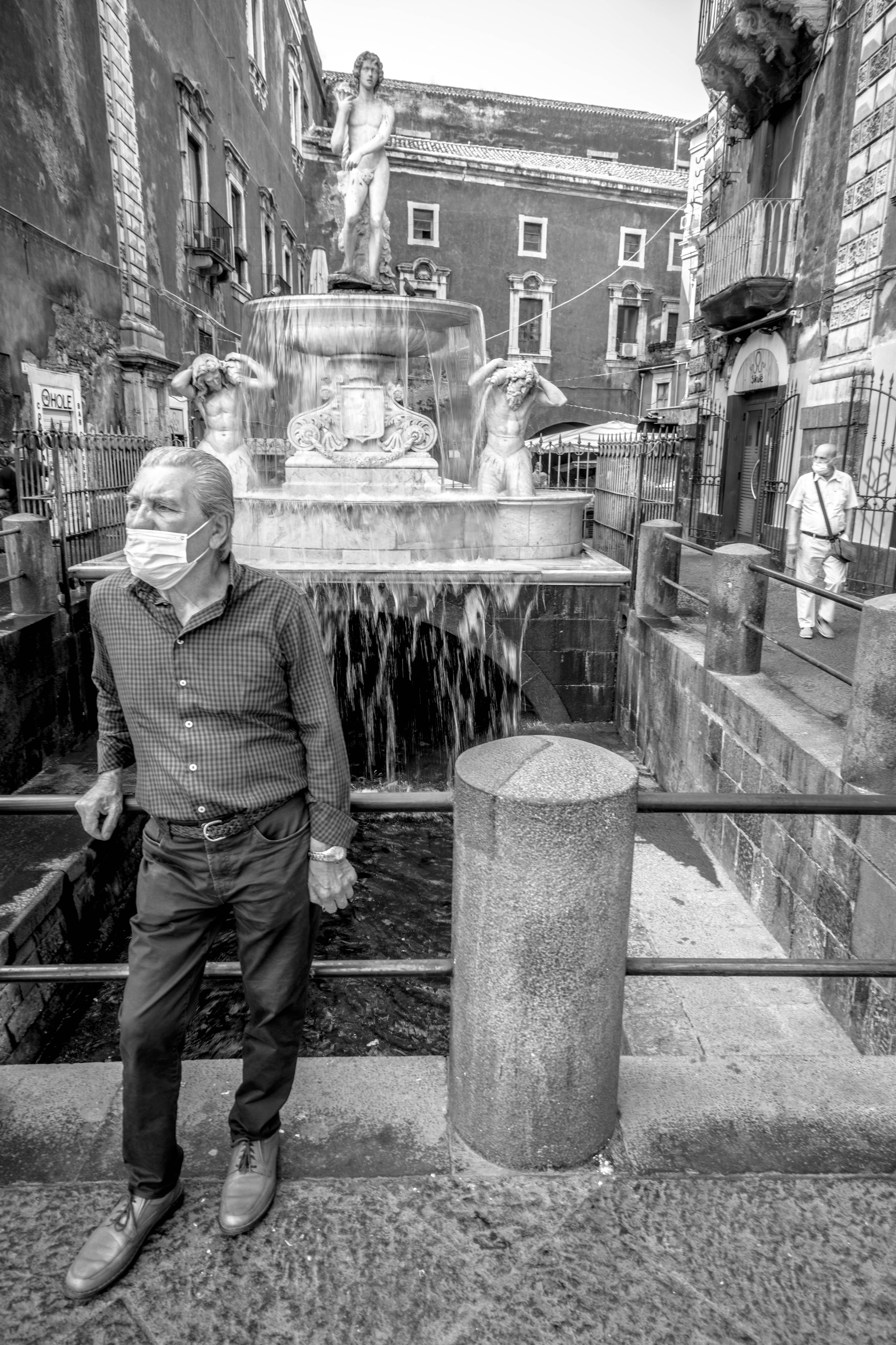 Masked local man standing before the Amenano Fountain in Catania, Sicily, in black and white