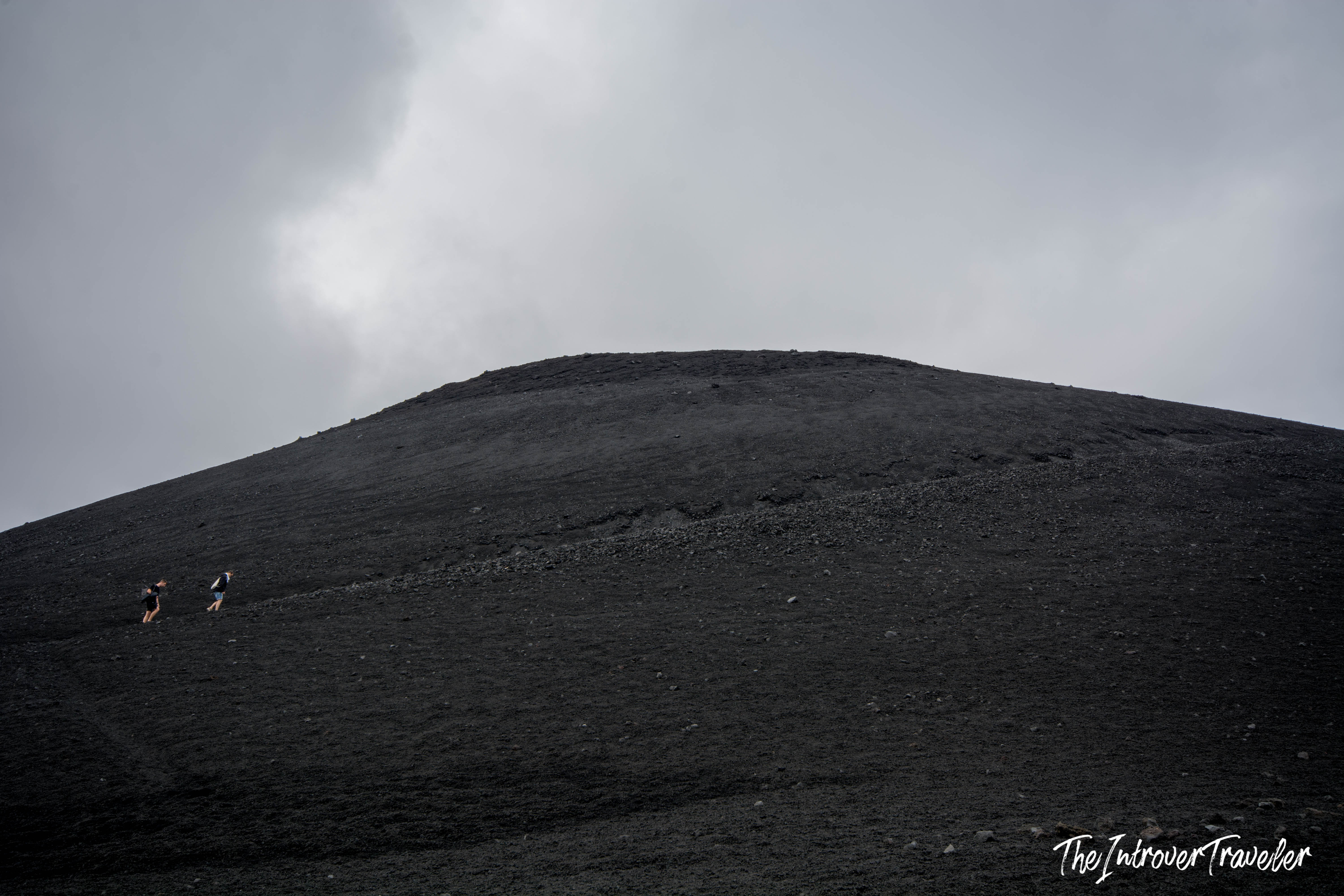 Two hikers ascending the vast black lava slopes of Mount Etna, Sicily, under a cloudy sky