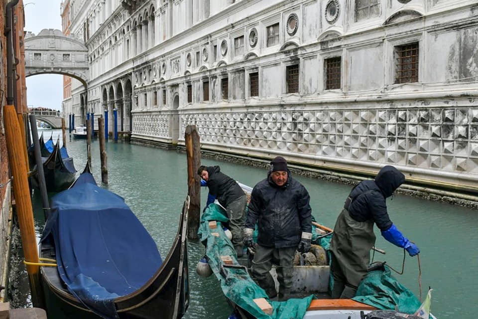 Workers on a boat dredging Venice canal beside the Doge's Palace, with Bridge of Sighs beyond
