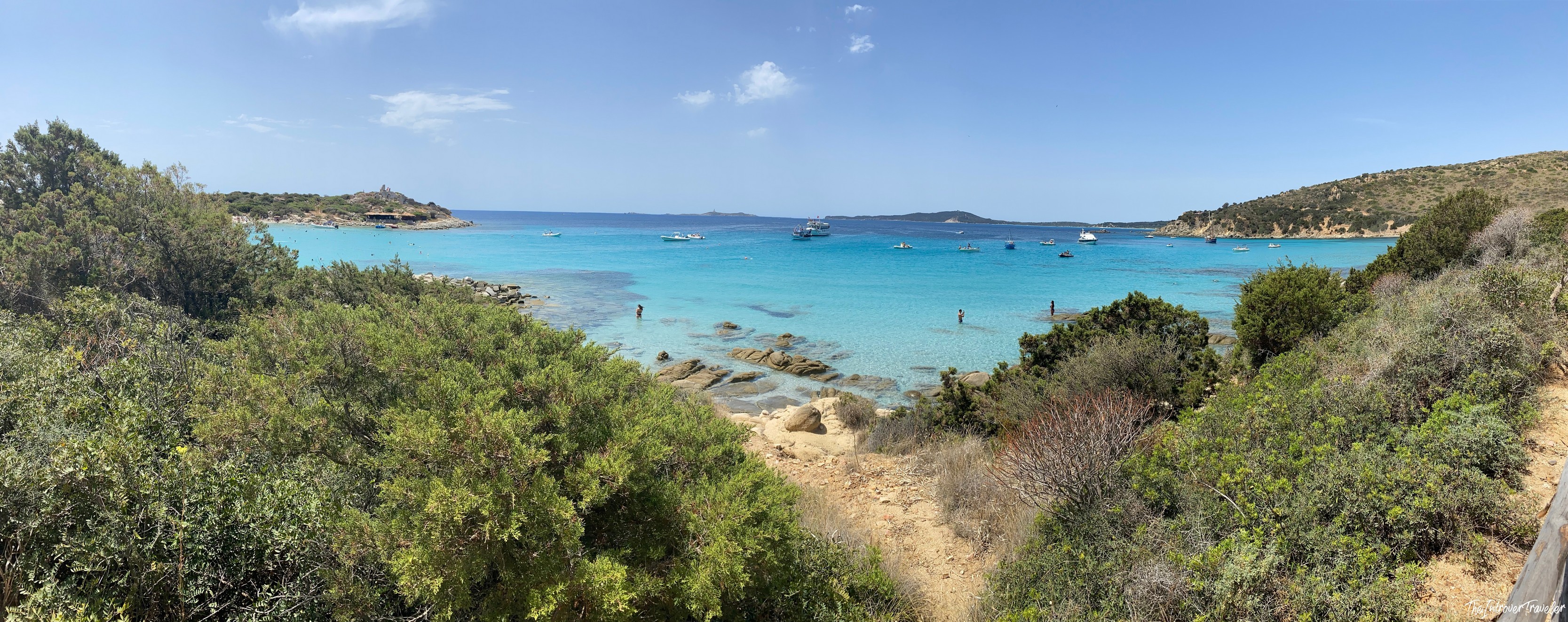Panoramic view of a turquoise cove in south Sardinia, with anchored boats, rocky shores, and Mediterranean scrub