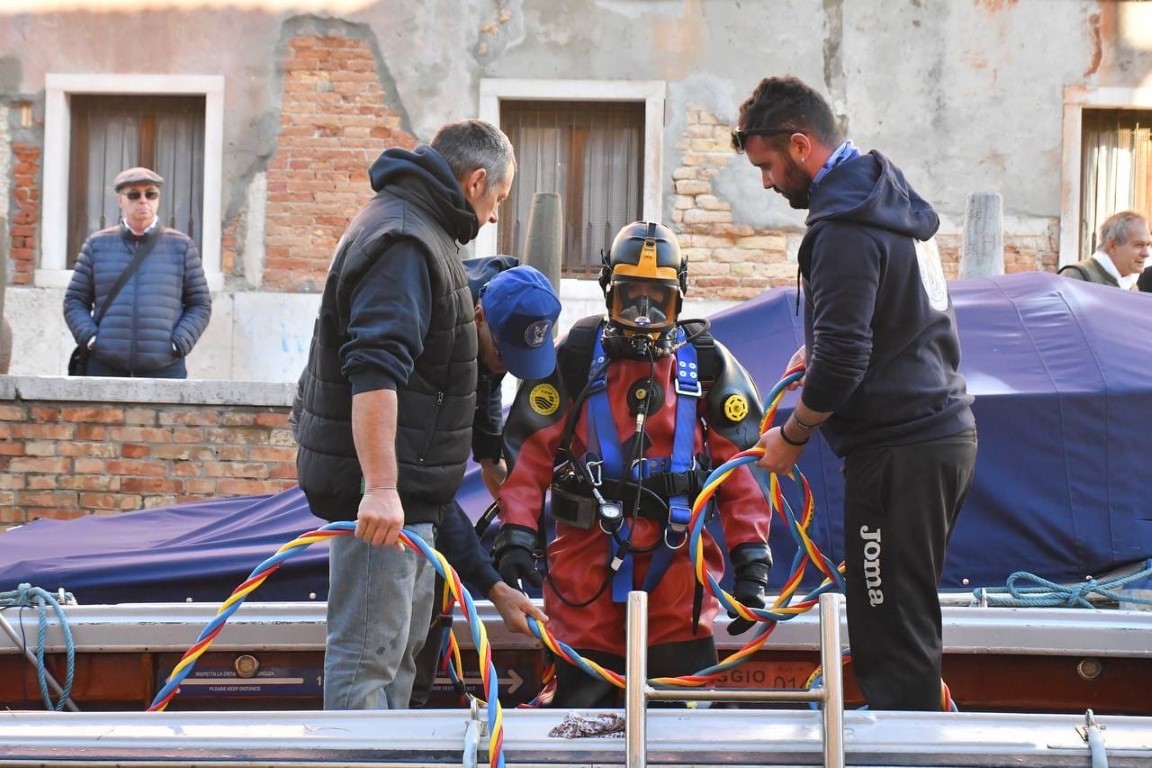 Venice canal diver in red dry suit helped by crew onto a boat along a Venetian waterway