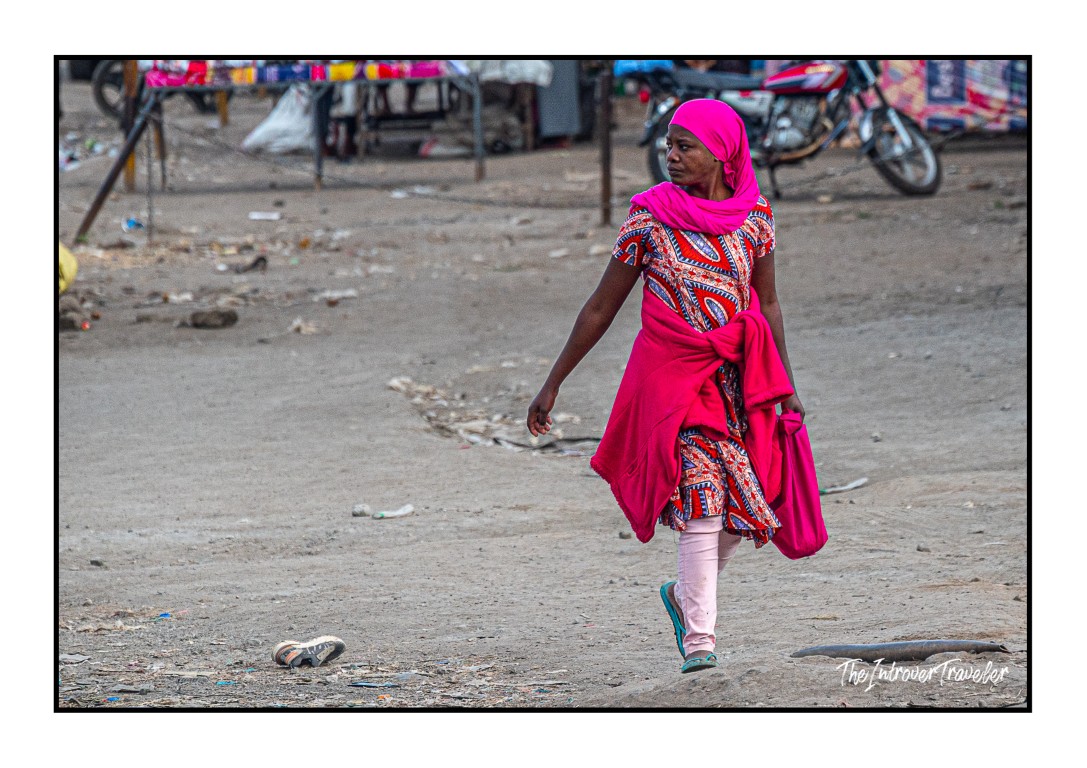 Woman in vibrant pink hijab and printed dress walking through a busy Tanzanian street market