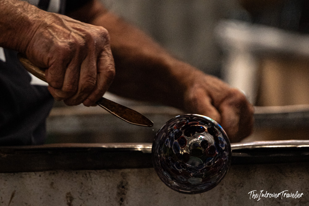 Murano glassmaker shaping a colorful molten glass piece with tools on a blowpipe in Venice