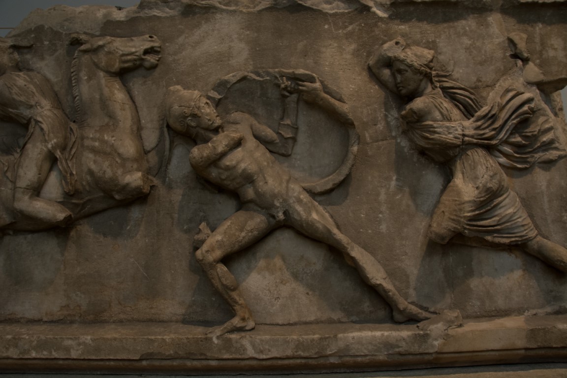 Ancient Greek marble frieze at the British Museum, showing an Amazon battle scene with draped female warrior and fallen male figure.
