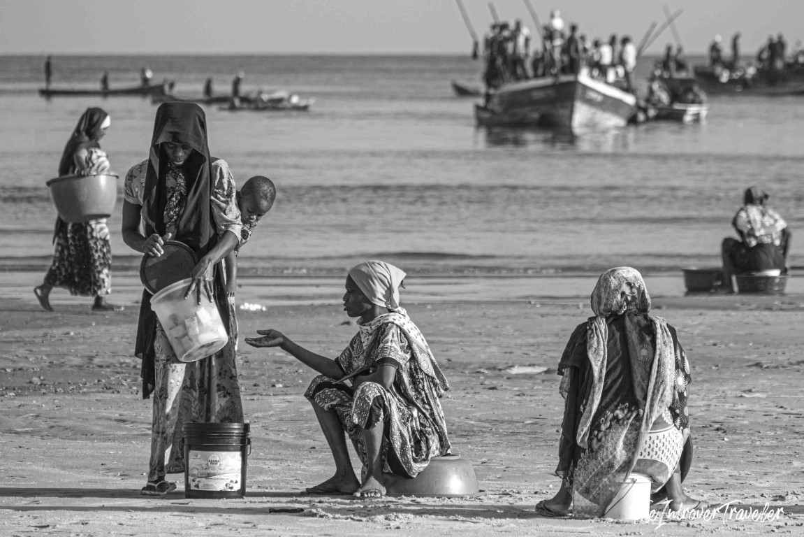 Women with buckets waiting on a sandy beach as fishing boats arrive at an African coastal fish market