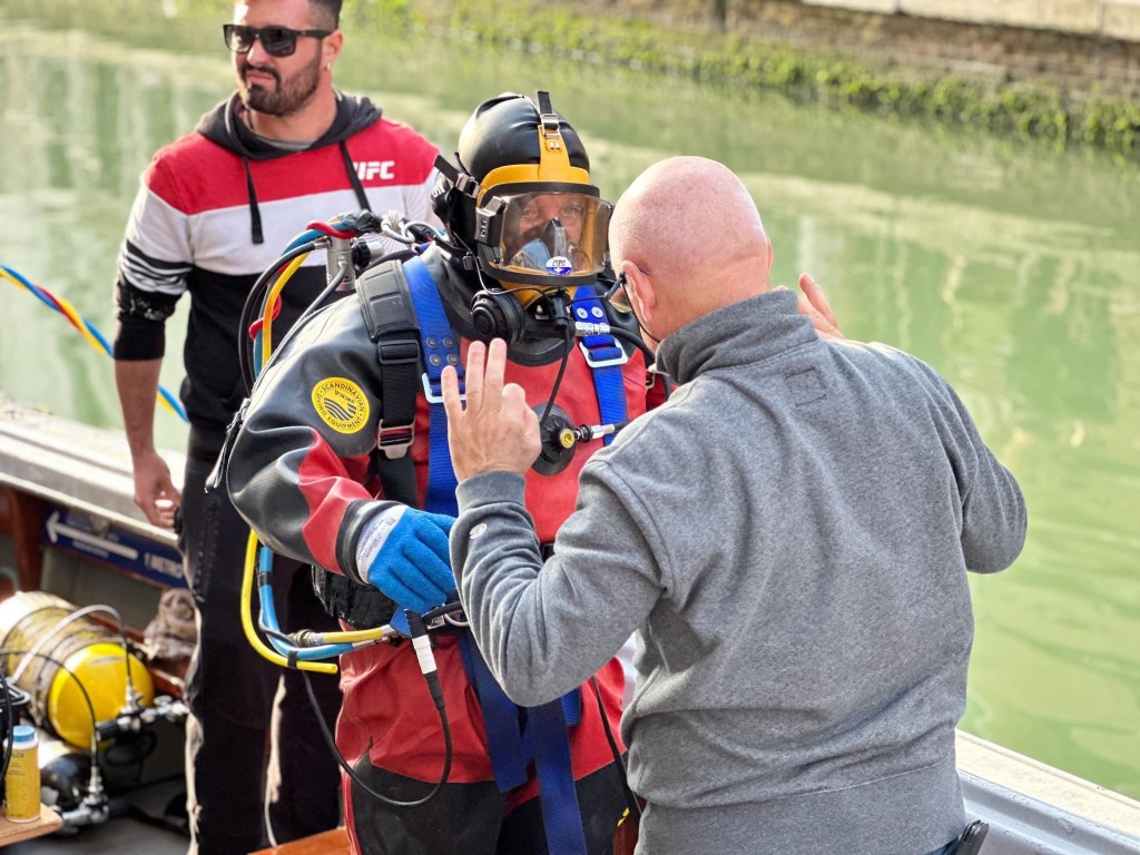 Diver in full dry suit and helmet being checked by crew on a Venice canal boat
