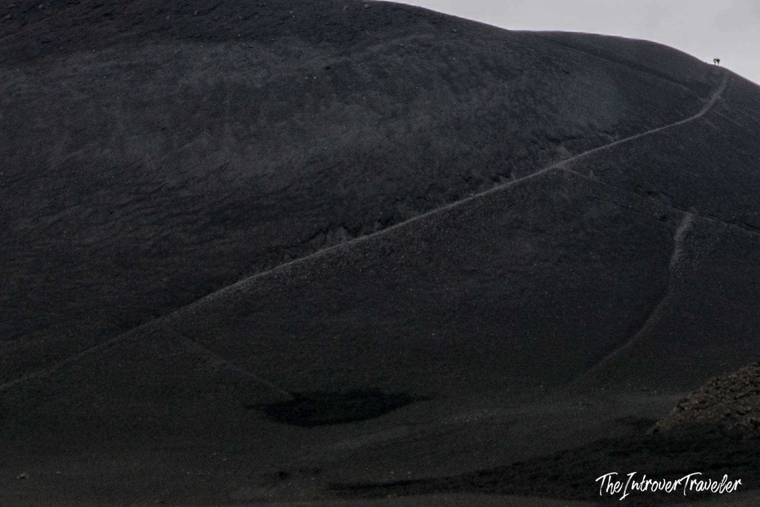 Black volcanic ash slopes of Mount Etna with a hiking trail and tiny figures at the summit ridge