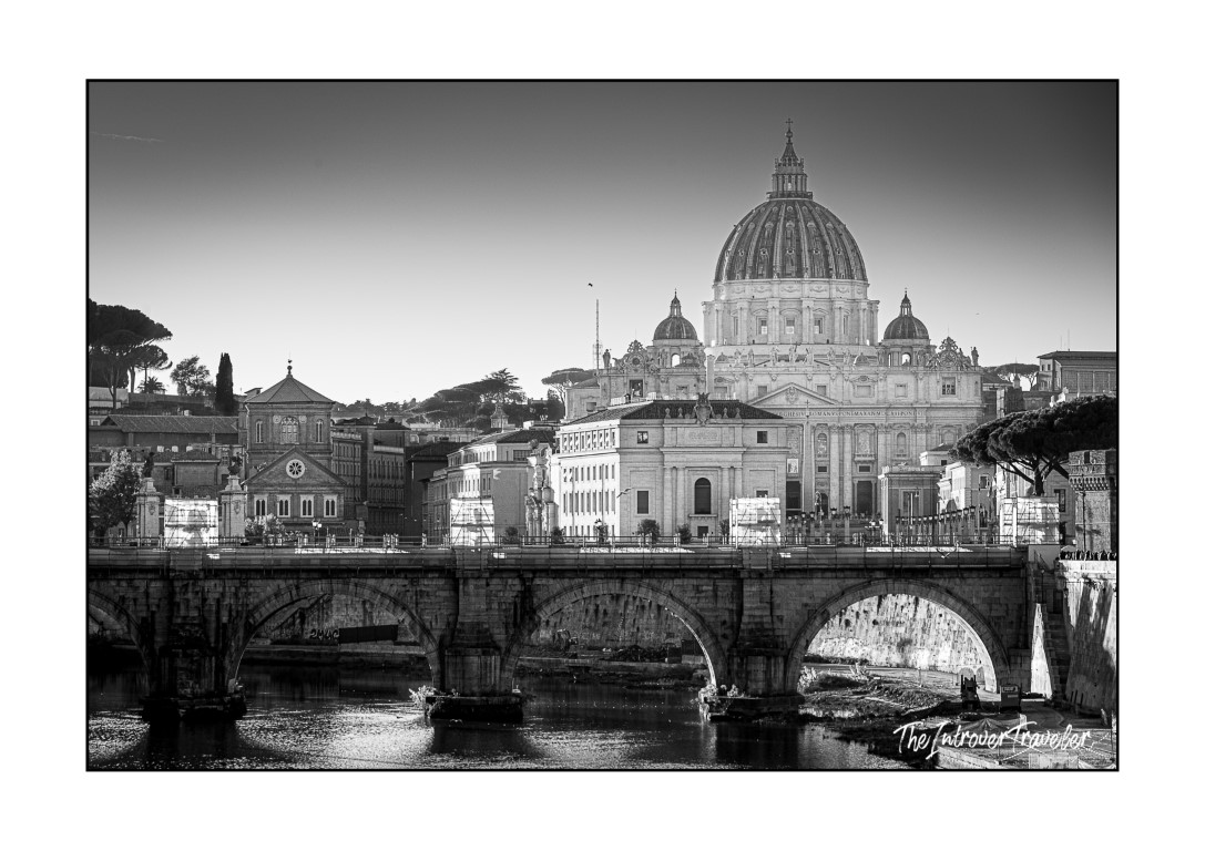 Black and white view of St. Peter's Basilica dome rising over Rome, seen across the Tiber and Ponte Sant'Angelo