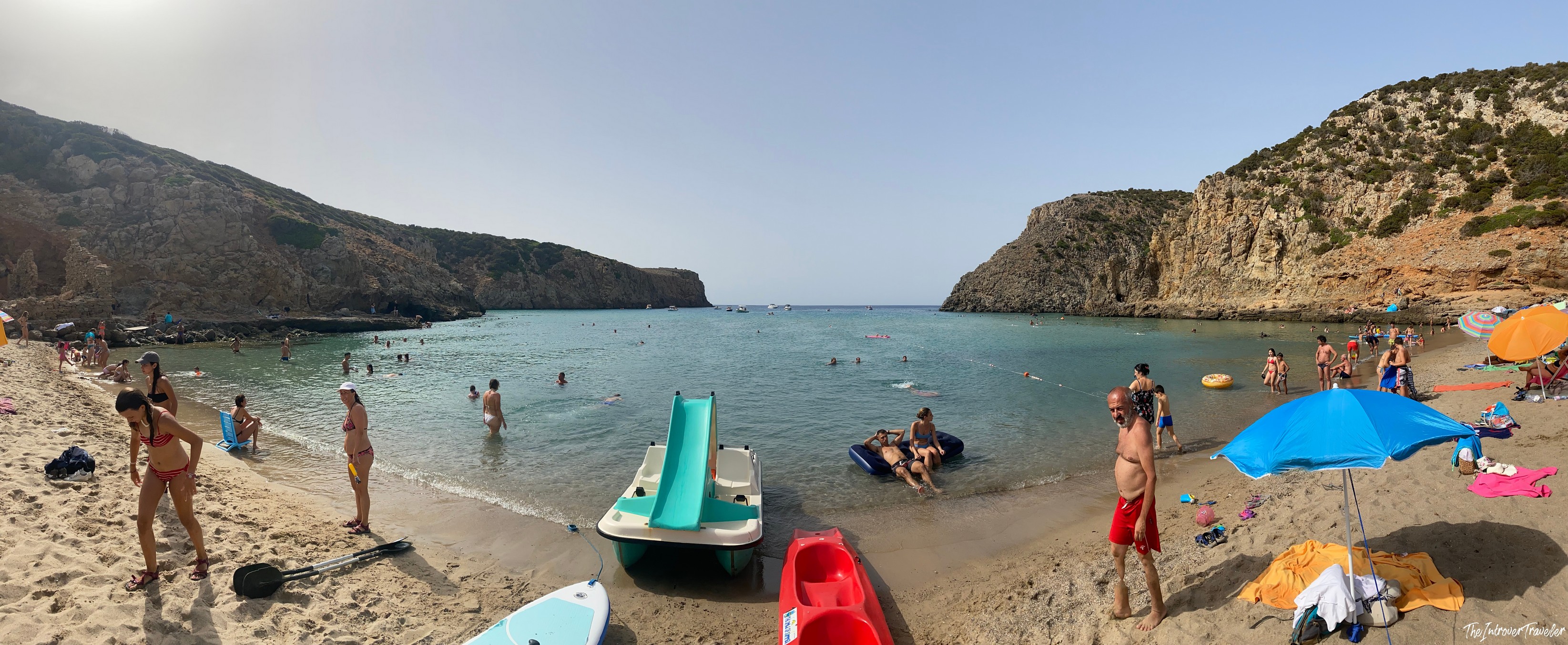 Busy sheltered cove beach in south Sardinia with turquoise water, rocky cliffs, and rental paddle boats