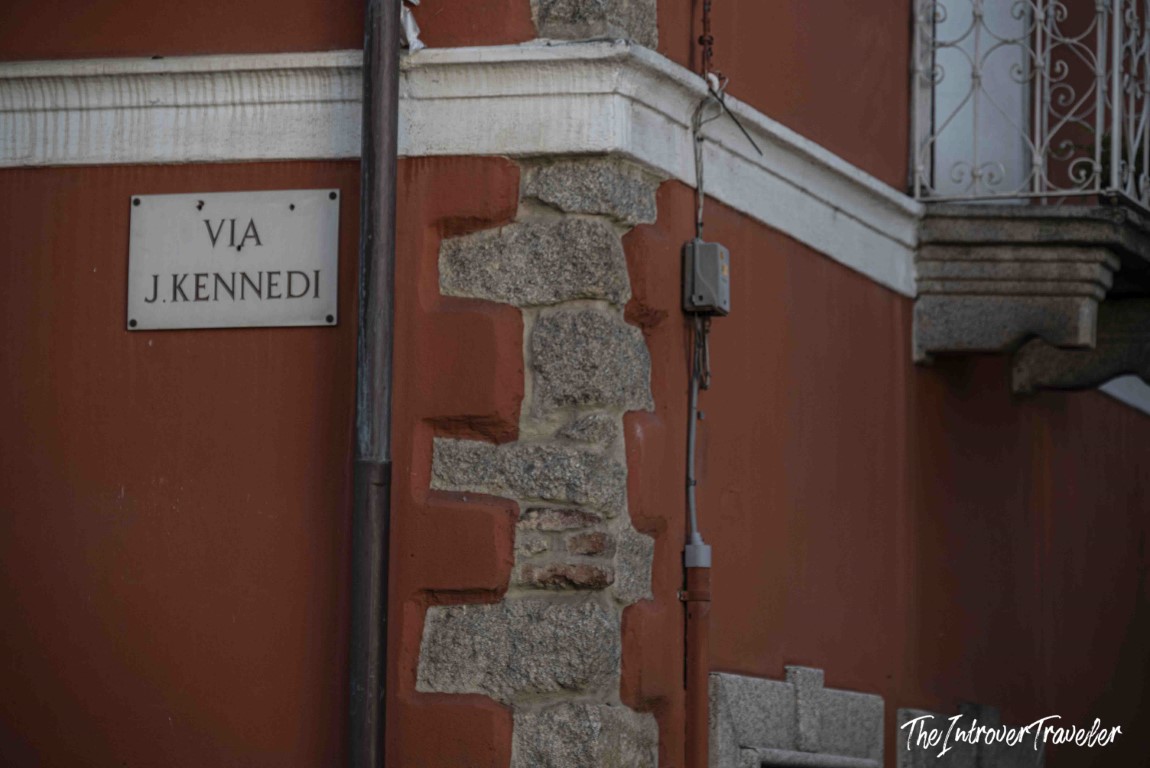 Street sign reading Via J. Kennedi on a red stone building in Orgosolo, Sardinia