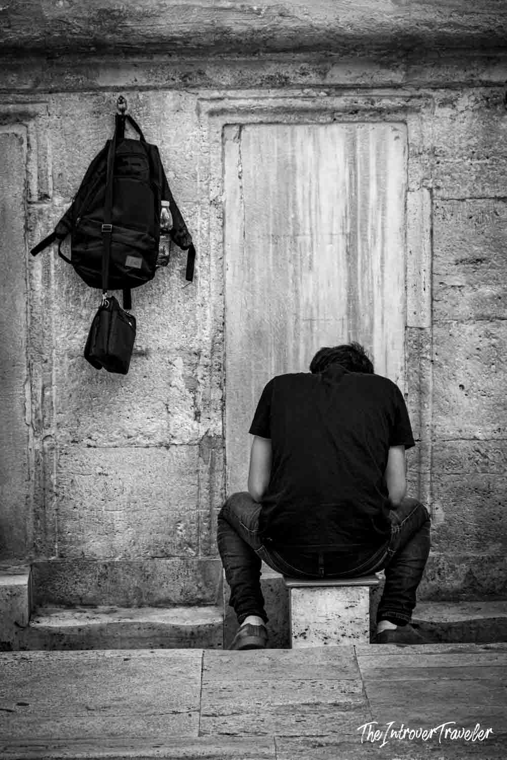 Solo traveler resting head down outside a historic Istanbul mosque stone wall with backpack hung nearby
