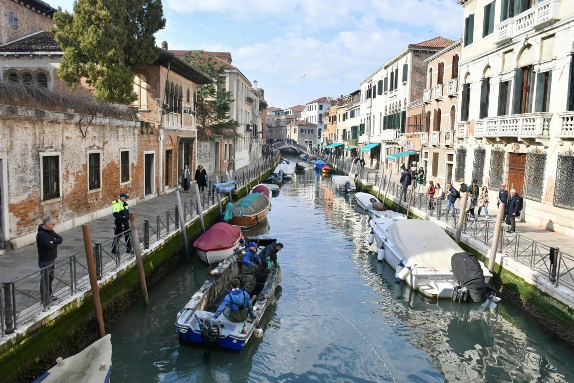 Venice canal workers in blue suits clearing debris from a narrow residential waterway lined with historic buildings