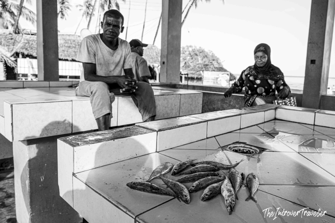 Vendors and fresh catch displayed on tiled counters at a coastal fish market near palm-fringed shores
