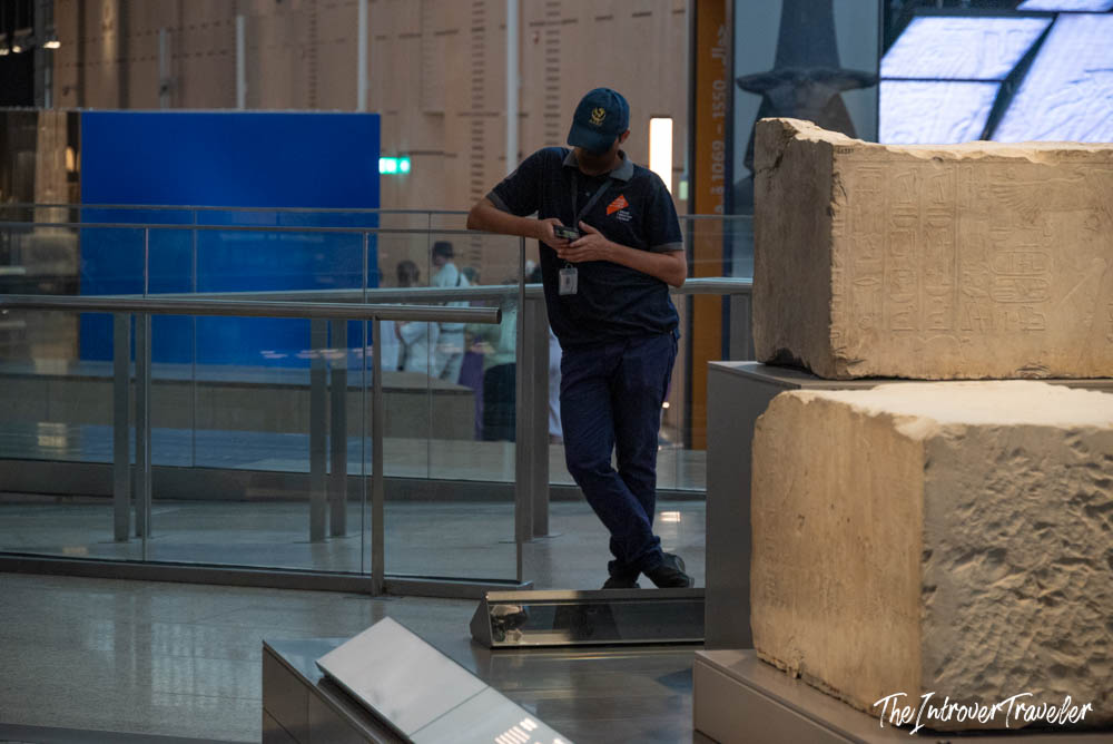 Museum guard checking phone beside ancient Egyptian stone blocks with hieroglyphs at the Grand Egyptian Museum, Cairo