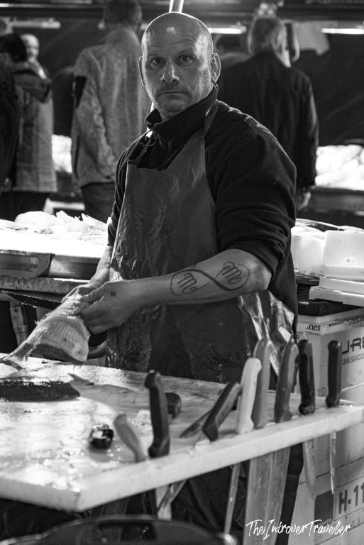 Tattooed fishmonger in apron handling fresh fish at a bustling coastal city fish market