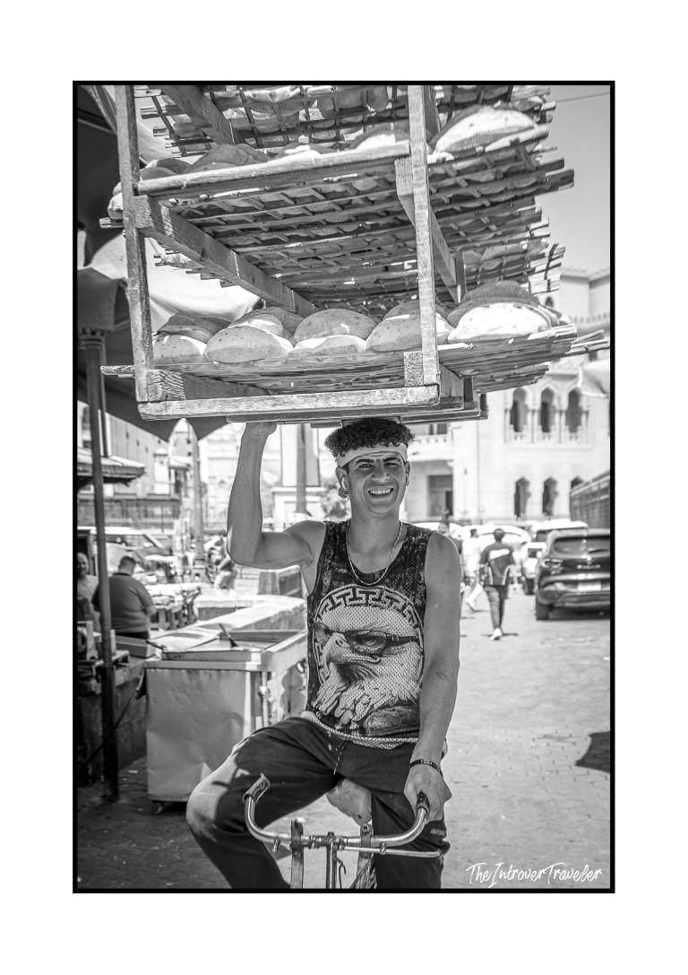 Young Egyptian bread seller balancing aish baladi loaves on his head while cycling through a Cairo street market