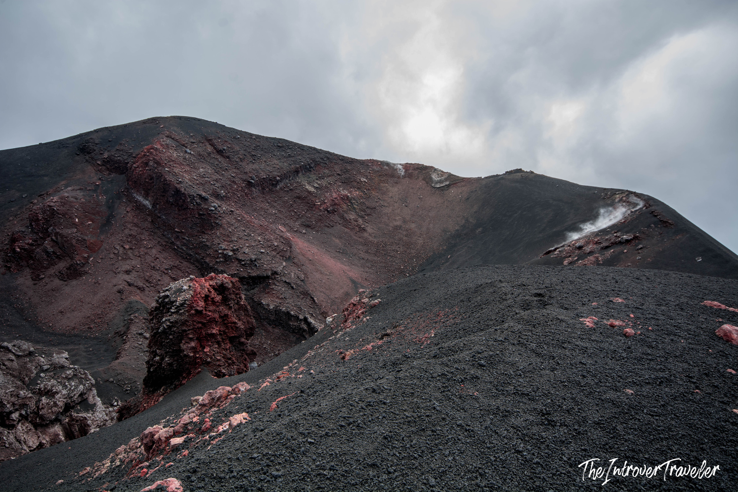 Mount Etna crater rim with black volcanic ash, red lava rock, and fumarole steam venting at the summit