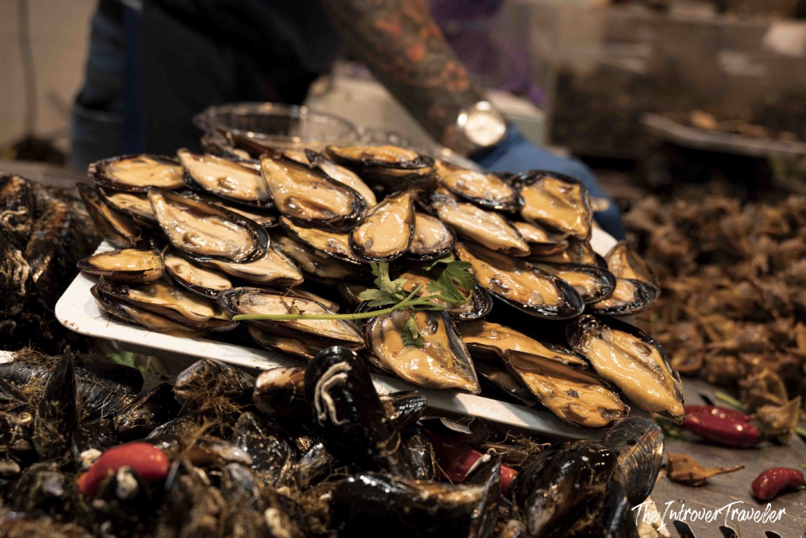 Piled fresh mussels on display at a bustling coastal fish market stall, garnished with chili and herbs