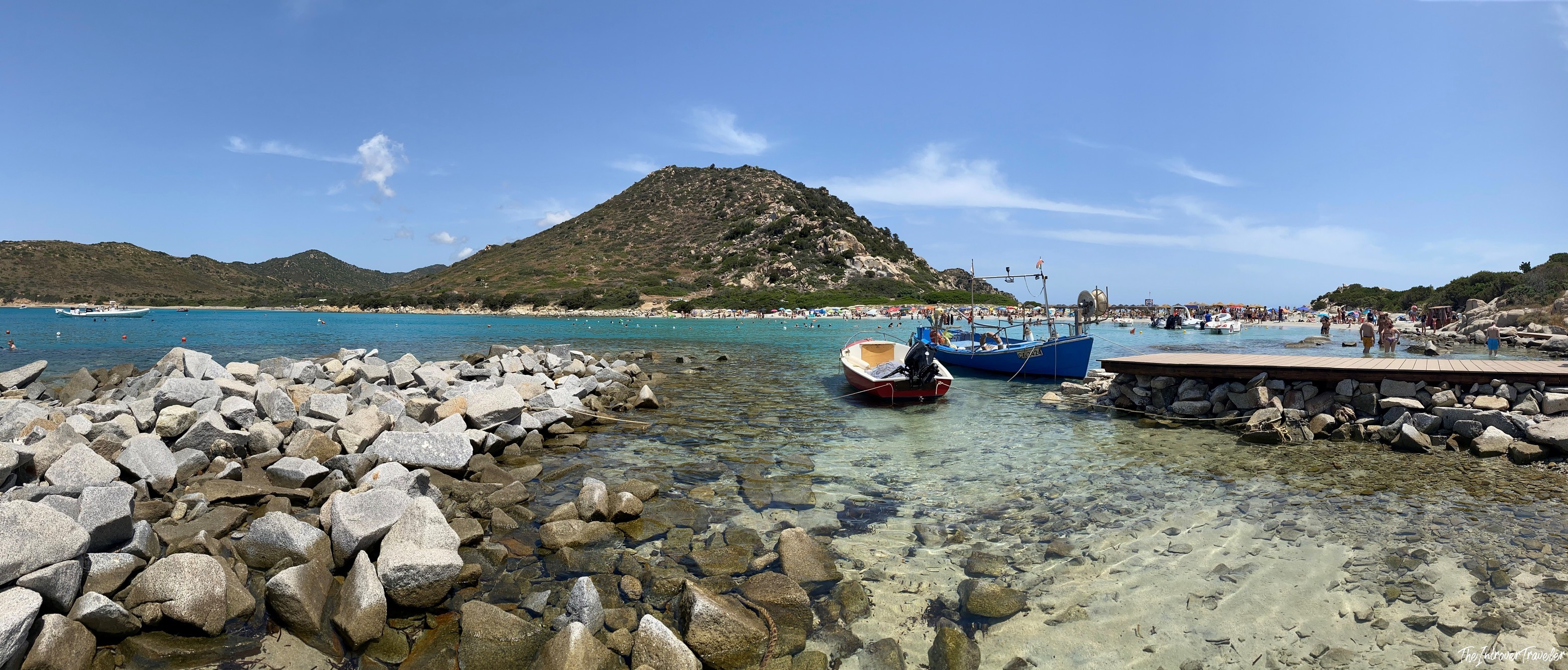 Small fishing boats moored at a rocky inlet near a south Sardinia beach, with turquoise water and a hill backdrop
