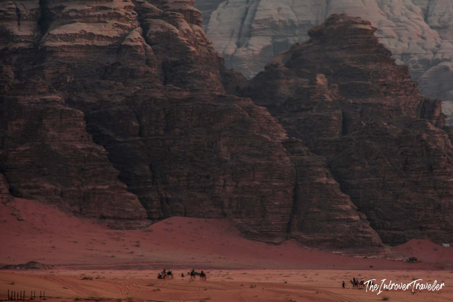 Camel riders crossing Wadi Rum desert, dwarfed by towering red sandstone cliffs in Jordan