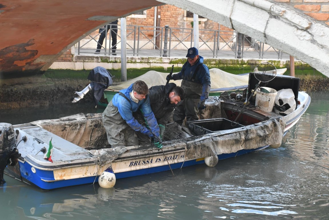 Workers on a mud-covered Brussa Is Boat vessel dredging a Venice canal beneath a bridge